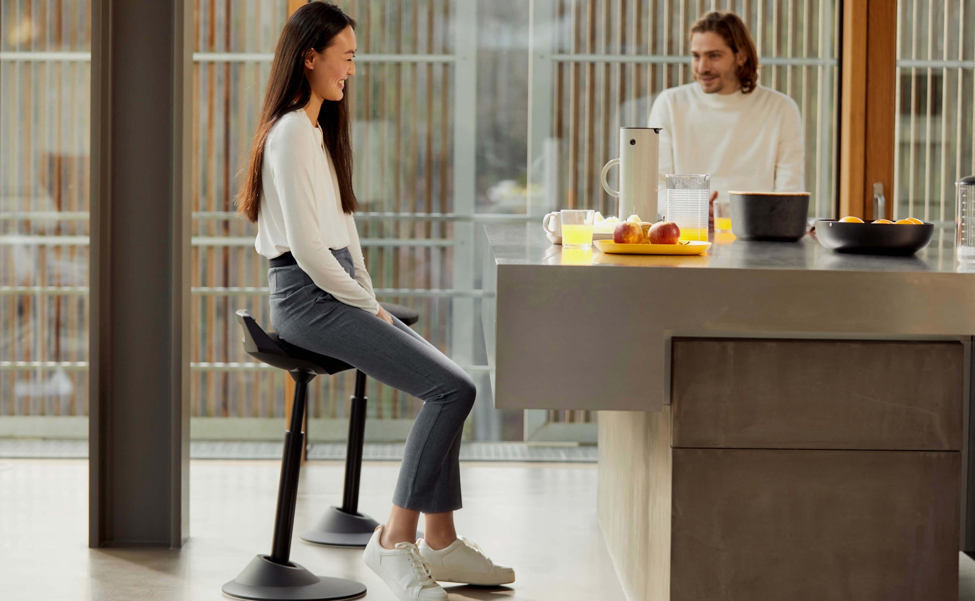 A woman laughs while sitting on sit to stand stool Aeris Muvman in the kitchen. Her friend facing her is making her laugh.