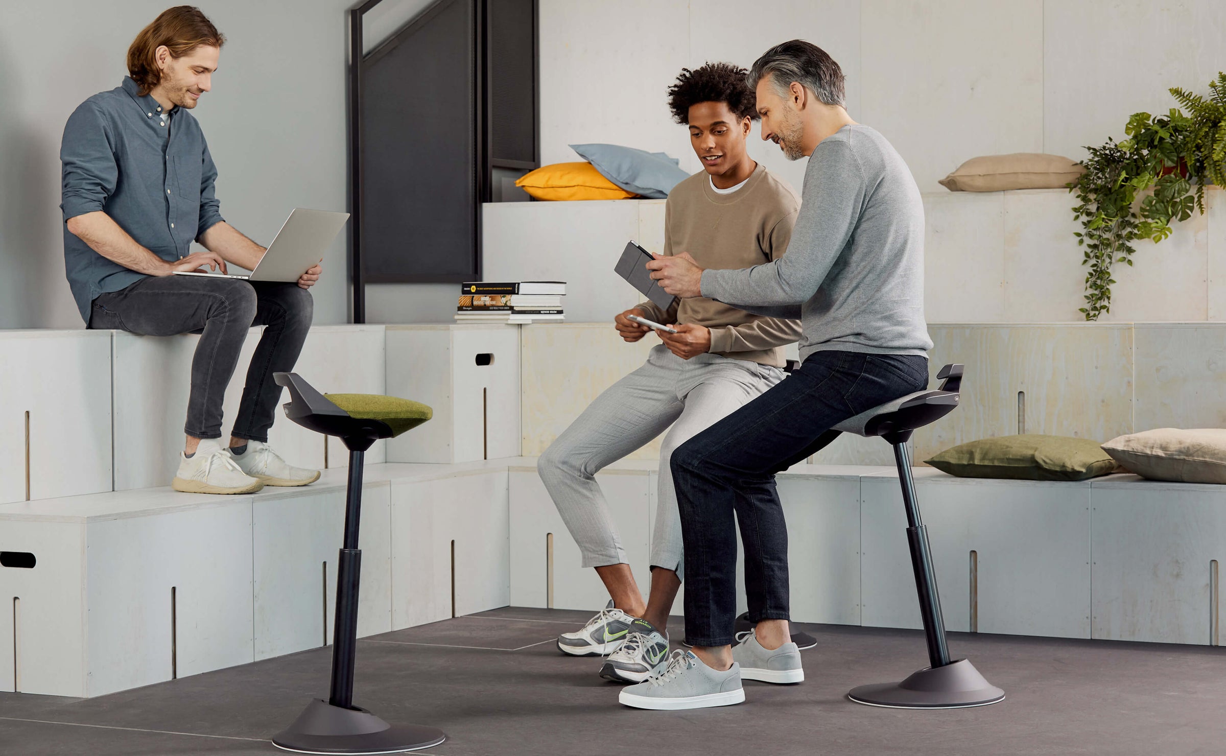 Three men having a meeting in an open work space. Two of them sitting on standing desk chair Aeris Muvman while discussing.