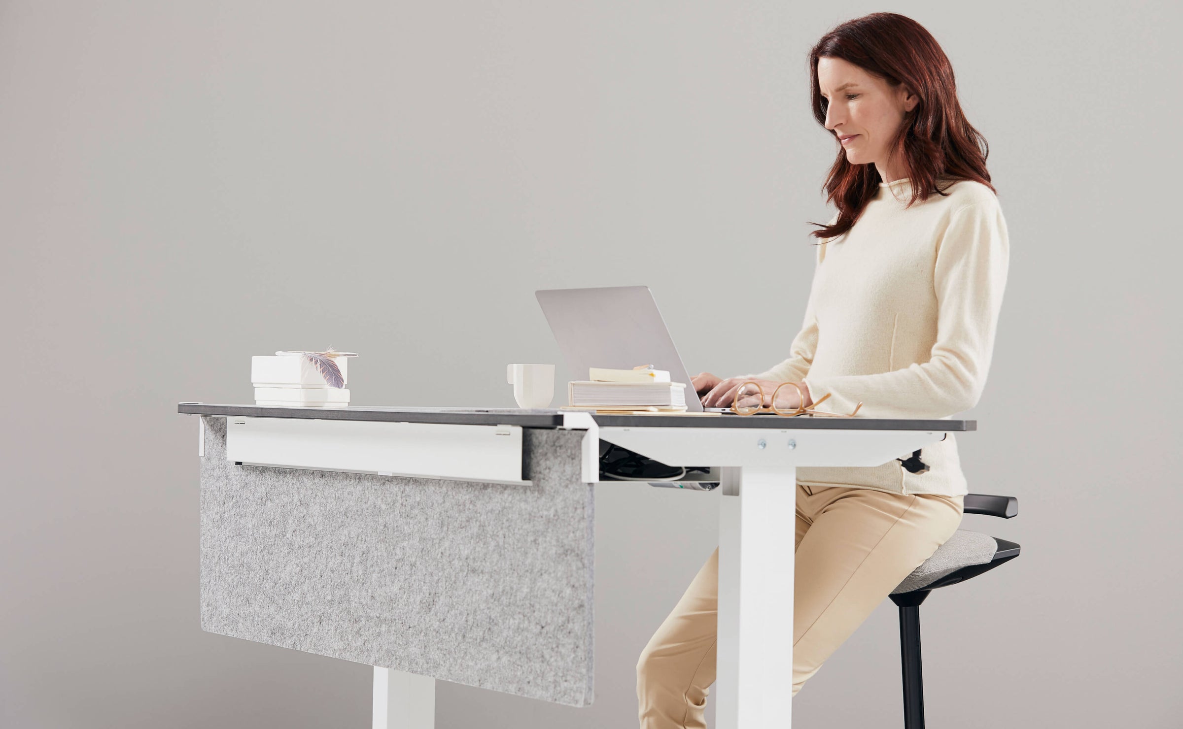 A woman is sitting on sit to stand stool Aeris Muvman while working on her laptop at her height adjustable desk.